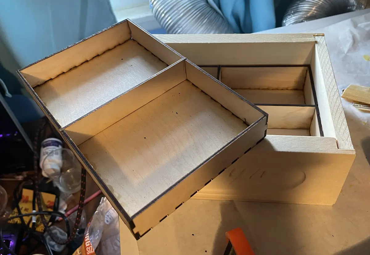 A photo shows two unfinished wooden boxes on a cluttered workbench; one box is open and tilted, revealing its empty…