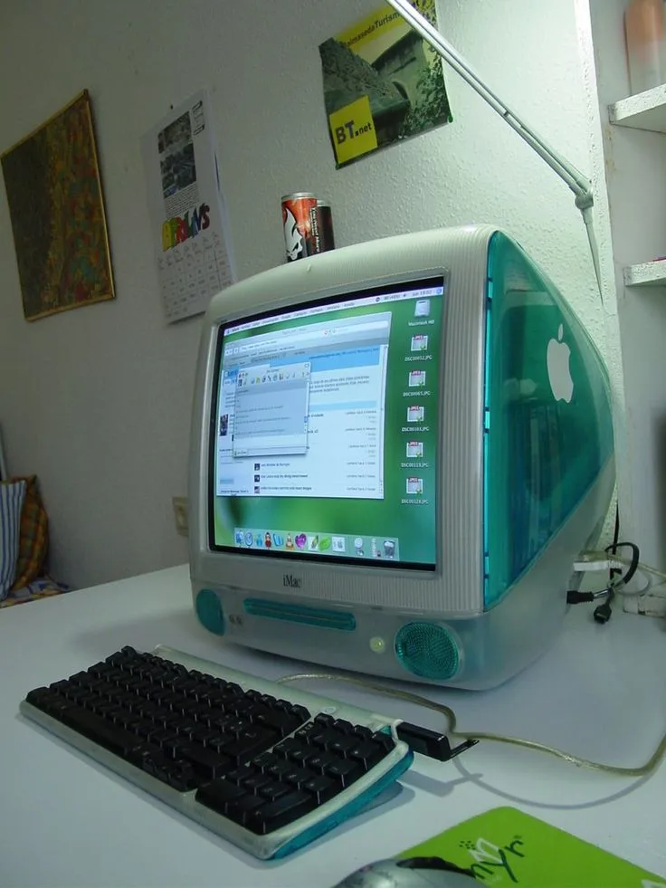 An old iMac G3 computer with a green translucent case and silver base sits on a white desk, its screen displaying a macOS…