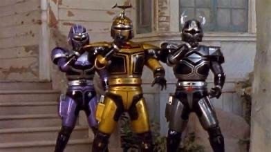 Three Power Rangers in purple, gold, and black armor stand side-by-side in a dilapidated building. The gold-armored Ranger…