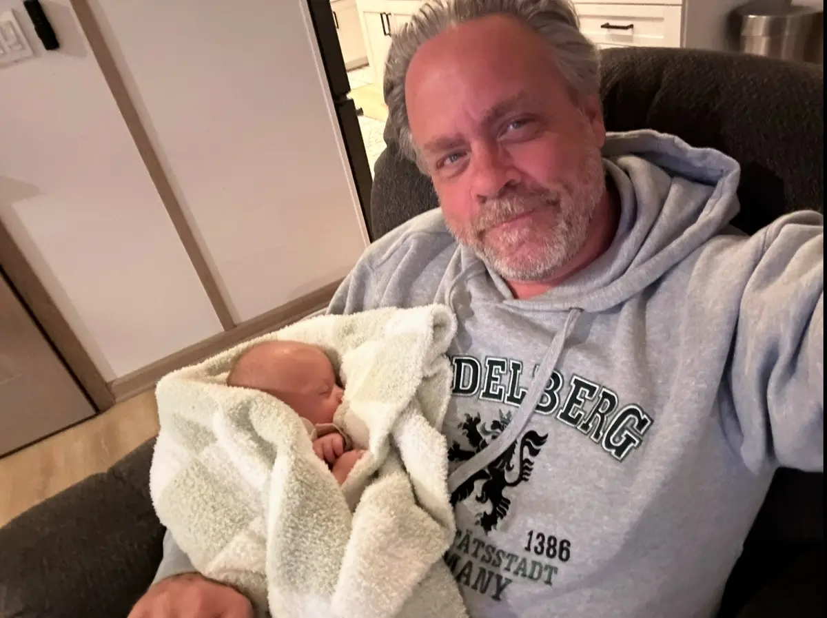 A middle-aged man with gray hair and a beard, wearing a gray hoodie, sits in a dark armchair holding a swaddled newborn…