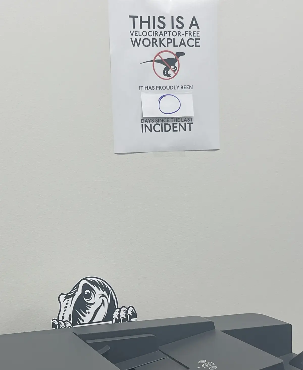 A white sign on a wall declares 'THIS IS A VELOCIRAPTOR-FREE WORKPLACE' with a crossed-out velociraptor icon, followed by…