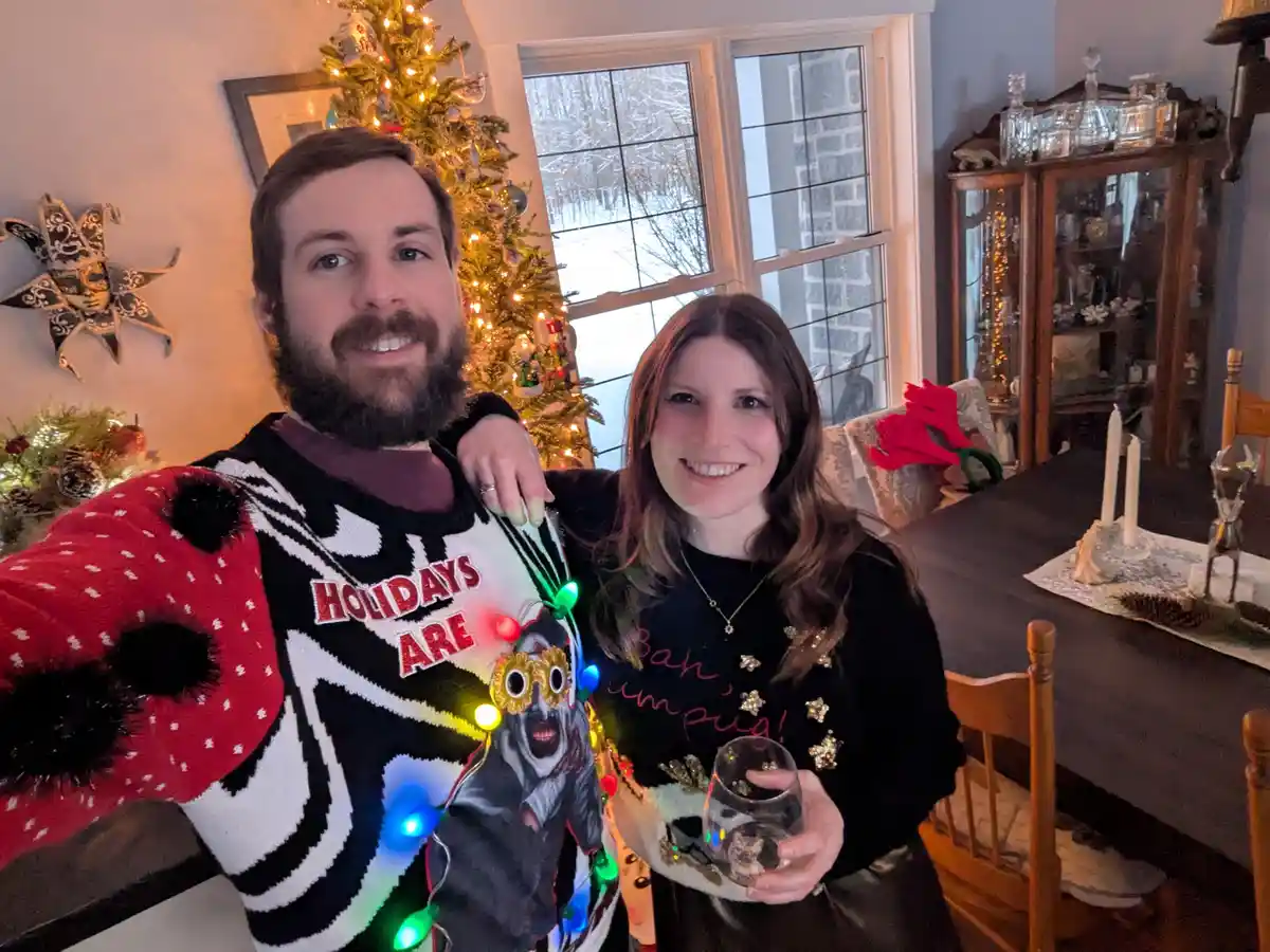 A man and woman pose for a selfie in a festively decorated room with a lit Christmas tree in the background. The man wears a…