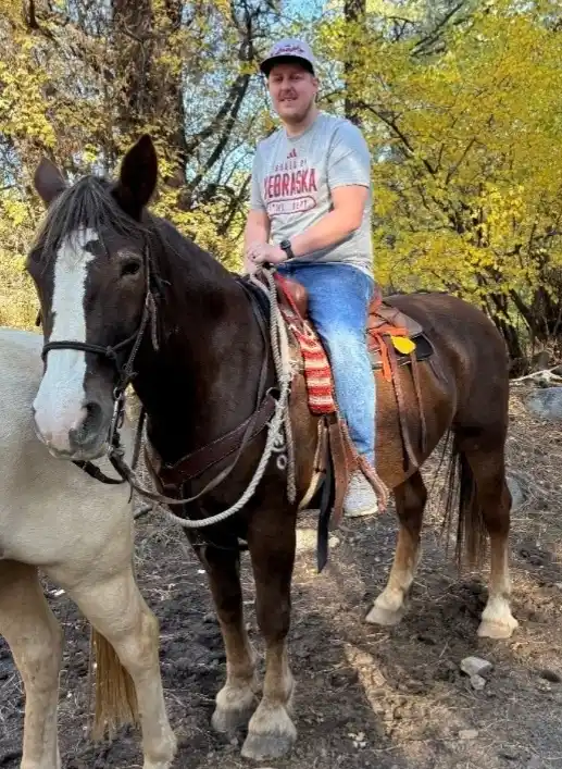 A man wearing a Nebraska t-shirt and cap sits atop a brown horse with a white blaze, holding reins. A second horse…