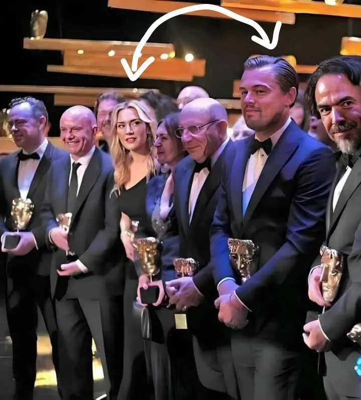 A group of people in formal black-tie attire stand on a stage holding golden BAFTA awards. A white arrow drawn over the…