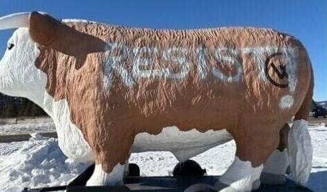 A large, life-sized statue of a brown and white cow stands in a snowy field under a clear blue sky. The word 'RESIST!' is…