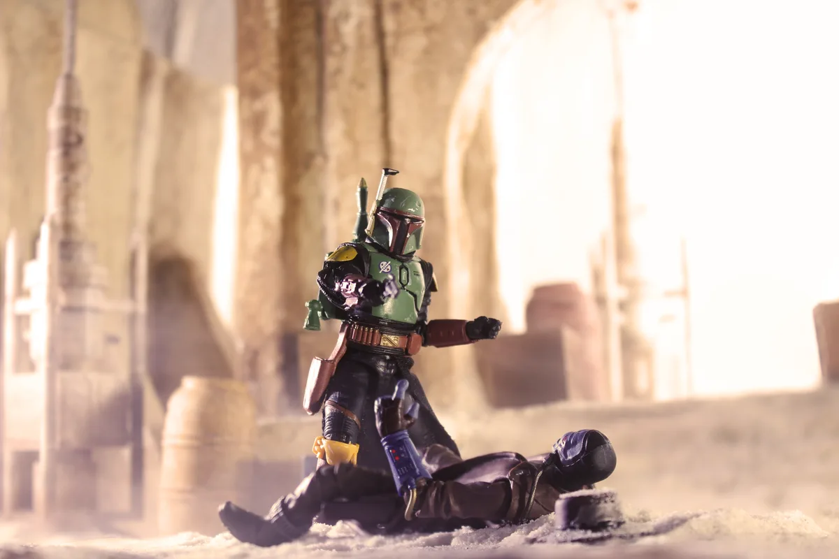 A Boba Fett action figure stands over a fallen action figure, likely a bounty hunter or assassin, in a desert-like setting…