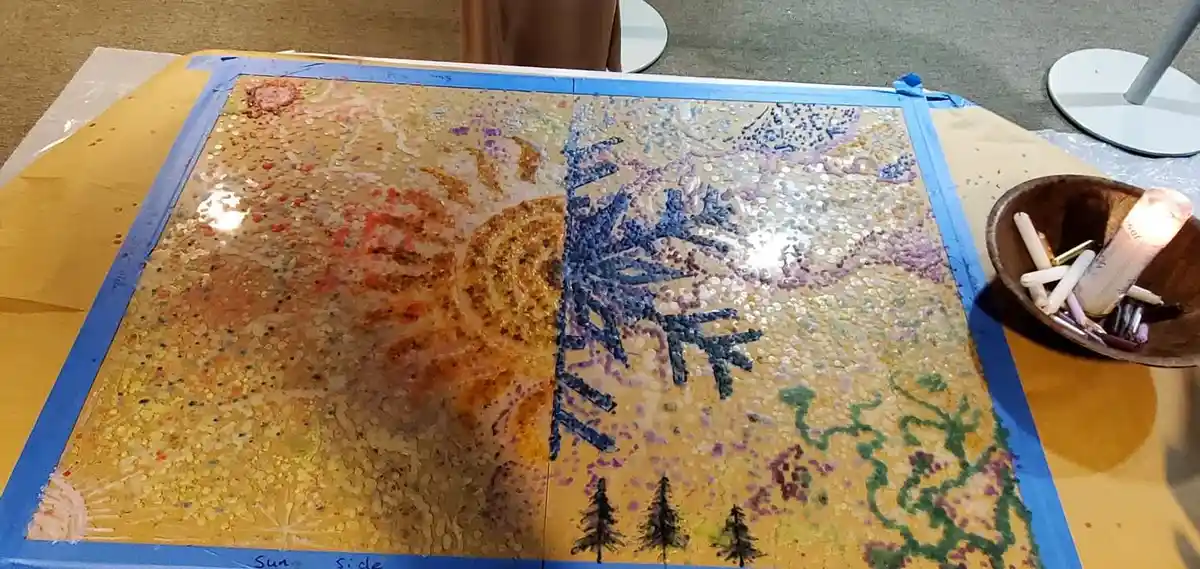 A large, square art piece taped to a table shows a split image: the left half depicts a sun with warm orange and yellow…