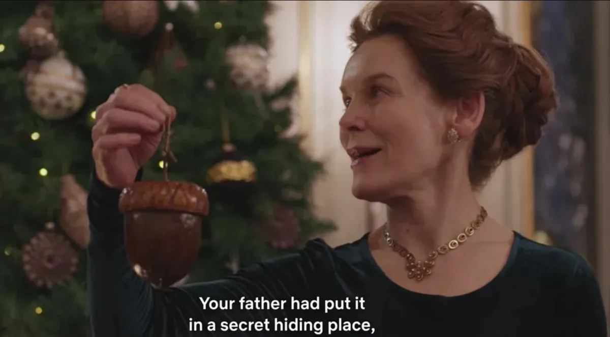A woman in a dark green velvet dress and gold jewelry holds up a large acorn ornament in front of a decorated Christmas…