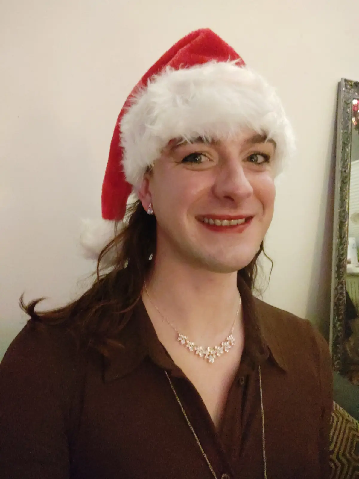 A person with long brown hair wearing a red and white Santa hat, a brown collared shirt, and a floral necklace smiles at the…