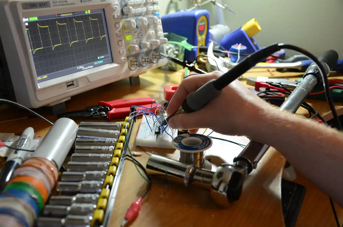 A person is soldering wires onto a breadboard in an electronics workshop, surrounded by tools like a soldering iron, spools…