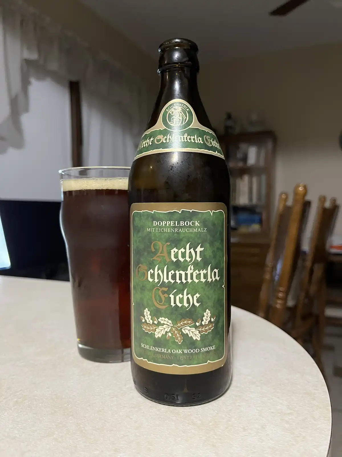 A dark brown bottle of Hecht Schlenkerla Eiche Doppelbock beer with a green label sits on a table next to a glass filled…