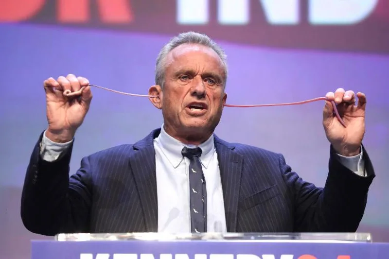 A man in a suit stands at a podium with 'KENNEDY' visible, holding a thin red string taut between his hands while looking…
