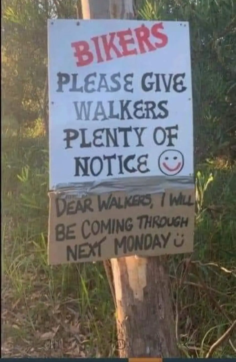 A handmade sign posted on a tree in a grassy outdoor area has two parts. The top white sign in red and black text reads…