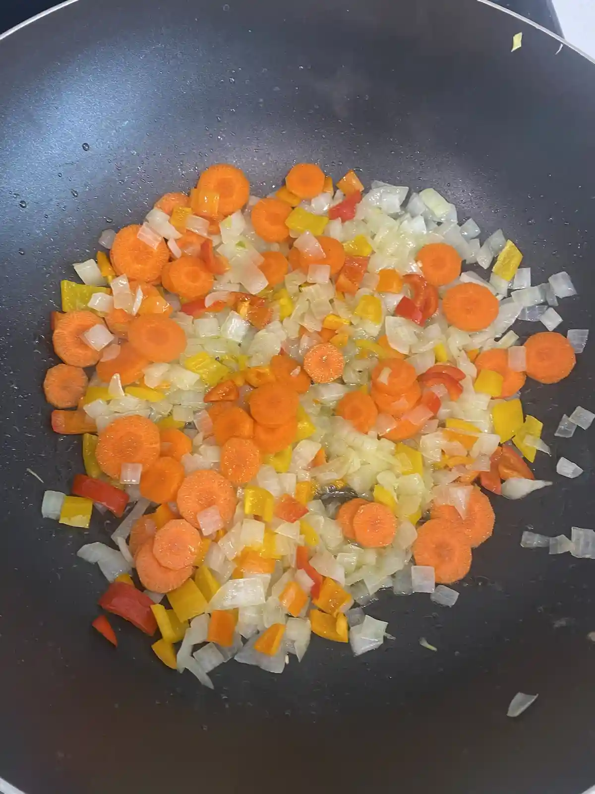 A close-up photo shows chopped vegetables—orange carrots, white onions, and red/yellow bell peppers—in a black frying pan…