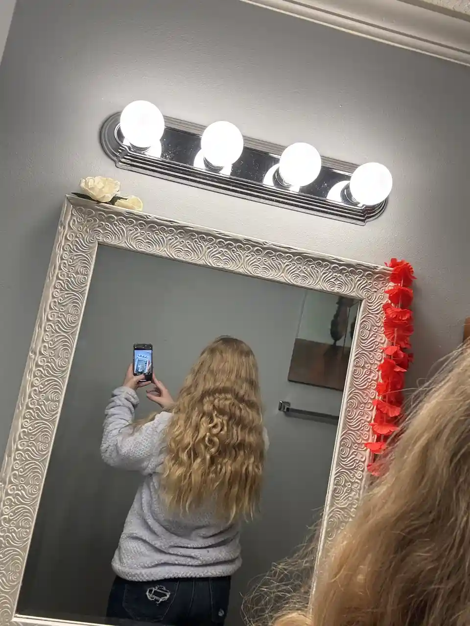 A person with long blonde hair, wearing a grey sweater and jeans, is taking a mirror selfie in a room with a large ornate…
