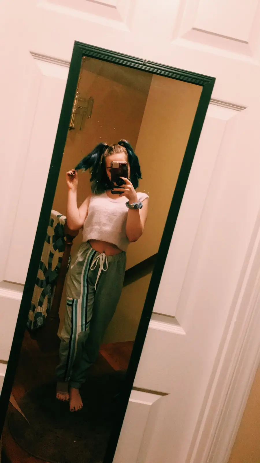 A person with blue-tipped pigtails takes a mirror selfie in a cropped white top and green track pants, holding a phone to…