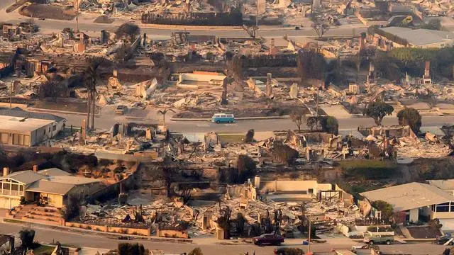 An aerial view shows a residential area devastated by fire, with most houses reduced to rubble and charred remains. A single…