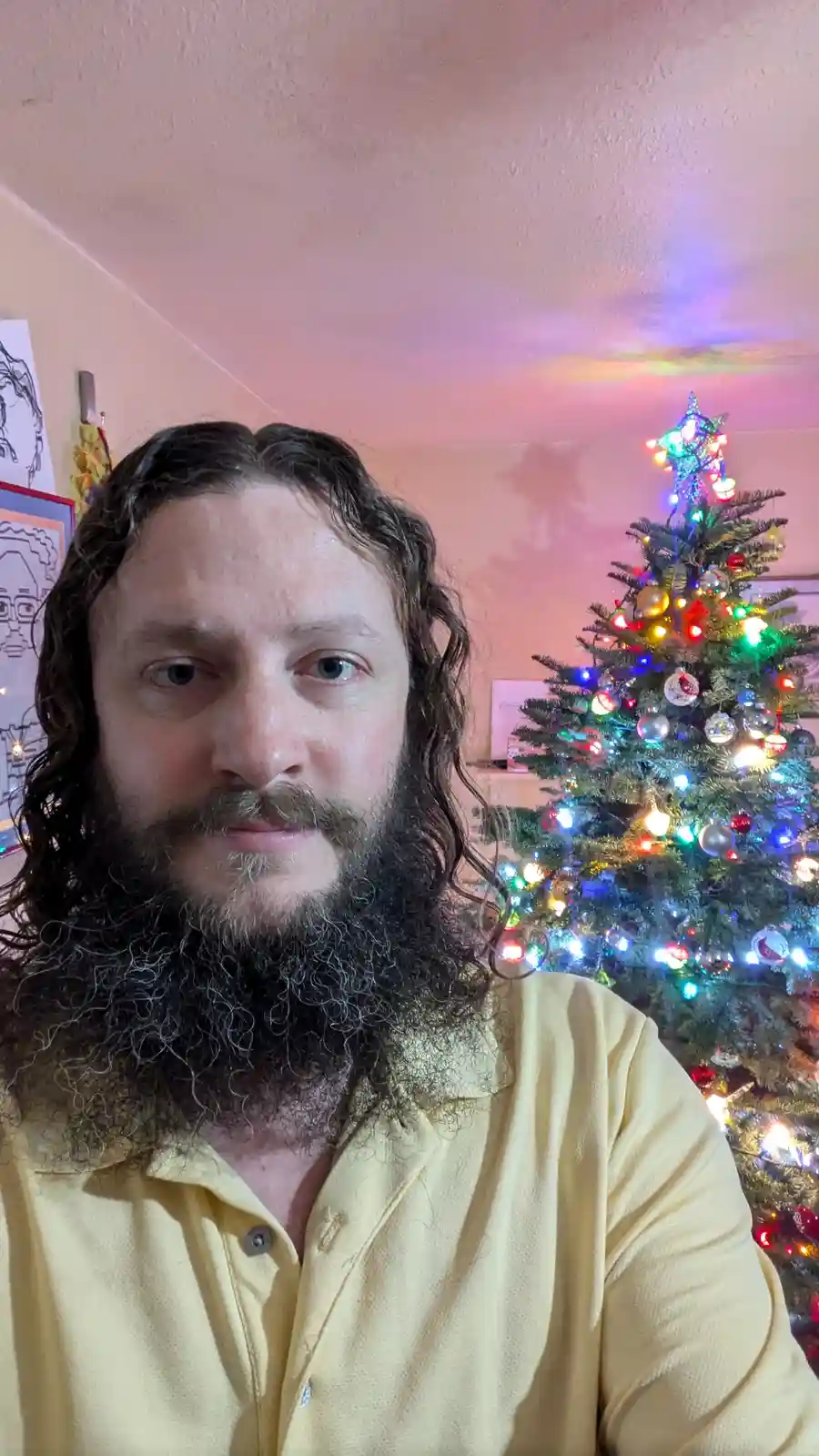 A man with long curly hair and a full beard, wearing a yellow polo shirt, stares directly at the camera with a neutral…