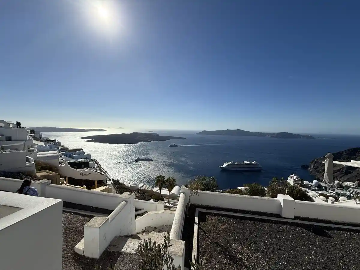 A scenic photo of a bright, sunny day in Santorini, Greece, showing white-washed buildings, a deep blue sea, and cruise…