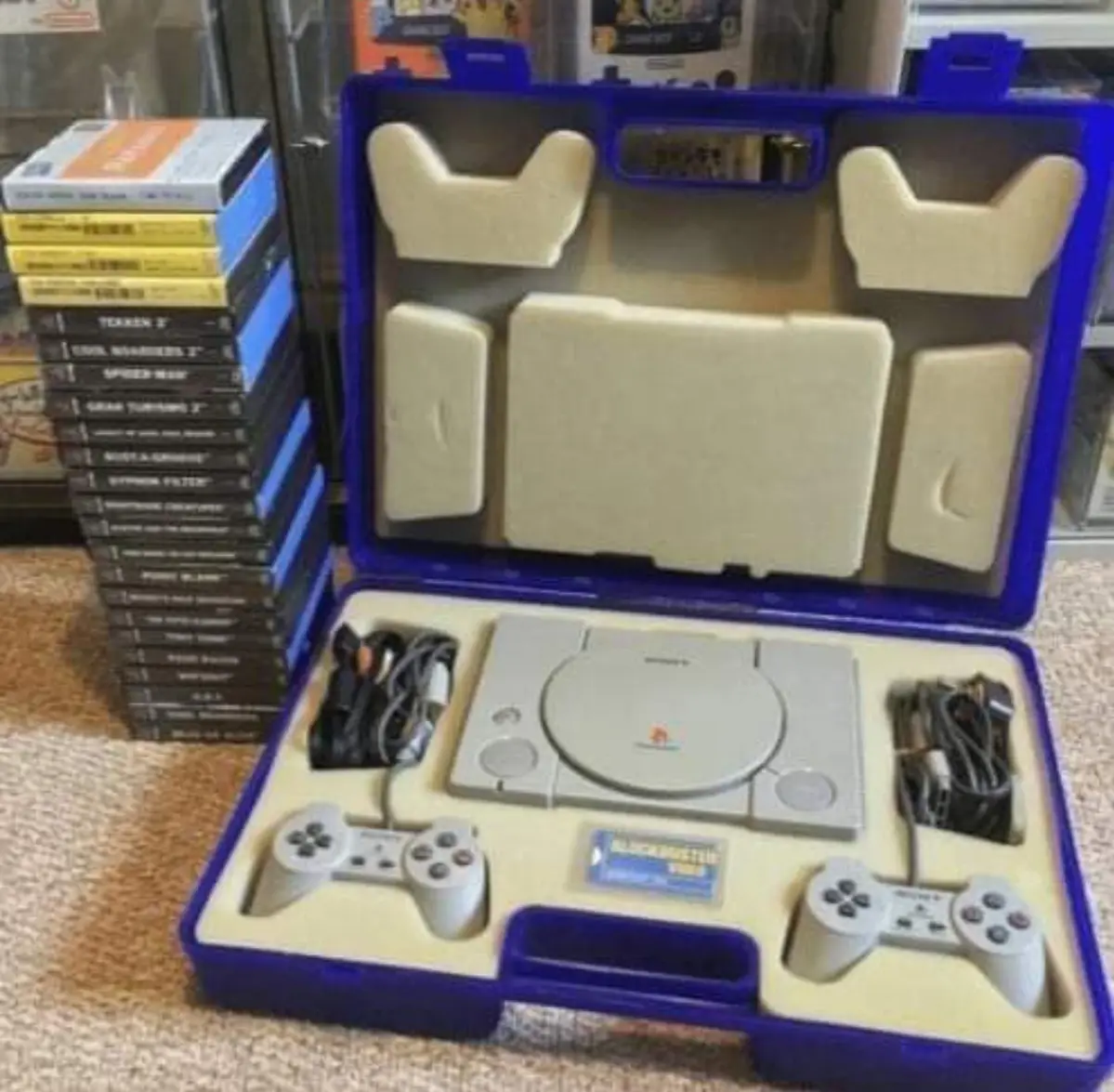 An open blue PlayStation 1 console case displays the grey console, two controllers, and cables nestled in beige foam. To the…