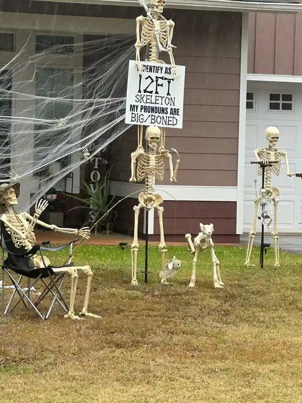 A Halloween yard scene with multiple plastic skeleton decorations. One tall skeleton holds a sign reading 'I IDENTIFY AS…