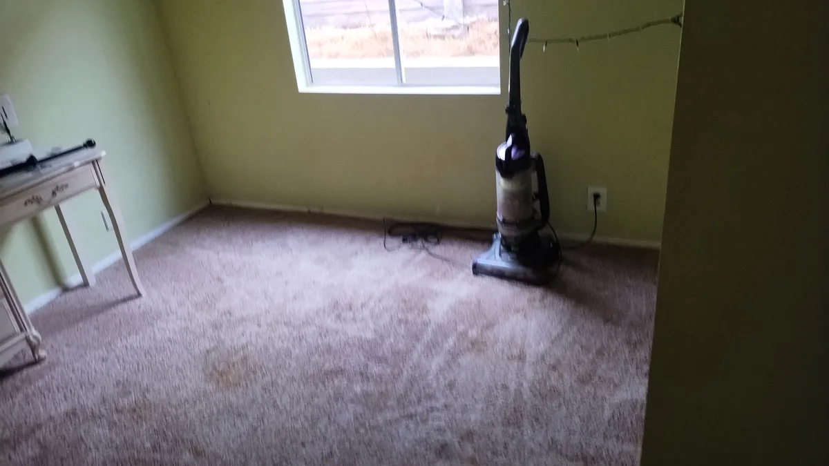 A messy, carpeted room with pale green walls, a window, and a vacuum cleaner standing upright near an outlet. A small white…