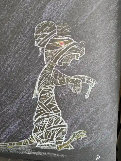 A chalk drawing on a dark gray surface depicts a cartoonish mummy with a single visible red eye, bared teeth, and a long…