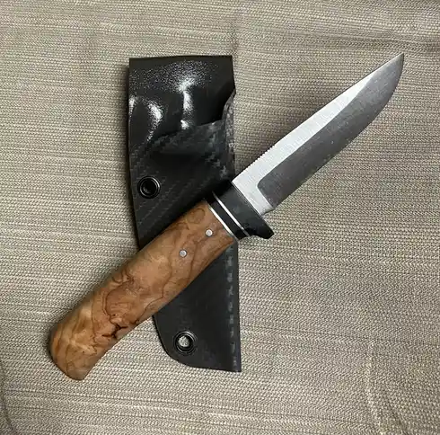A custom-made knife with a wooden handle and a serrated blade rests beside its black carbon fiber sheath on a textured beige…