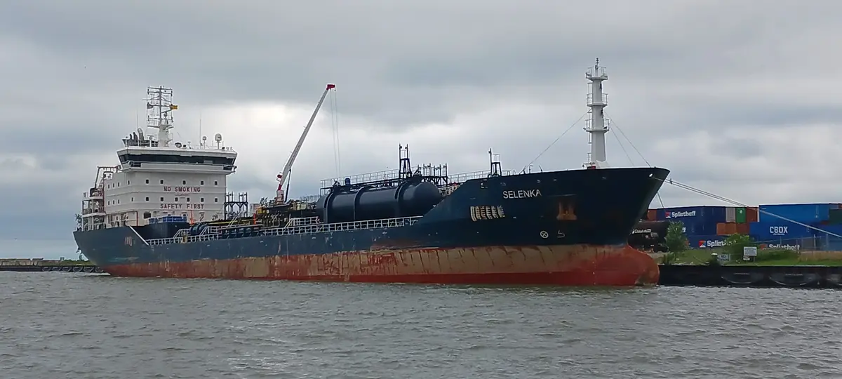 A large blue and red cargo ship named 'SELENKA' is docked in a harbor under an overcast sky. The ship's white bridge has 'NO…