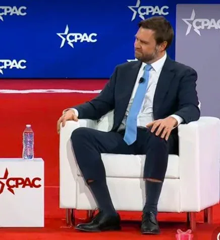 A man in a dark suit and light blue tie sits in a white armchair on a stage with 'CPAC' logos in the background. He is…