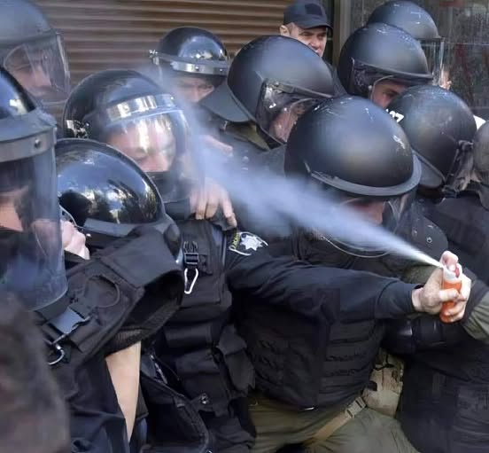 A group of police officers in riot gear are huddled together, one officer in the foreground is spraying pepper spray…