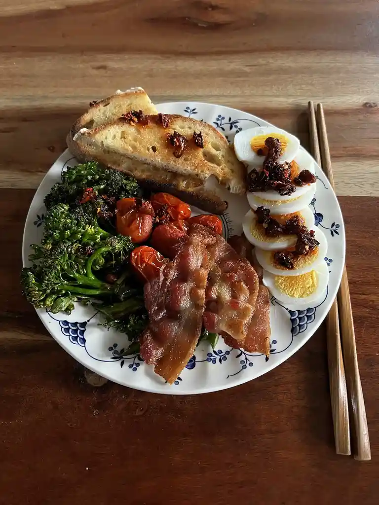 A plate of food on a wooden table includes roasted broccoli, cherry tomatoes, crispy bacon, sliced hard-boiled eggs topped…