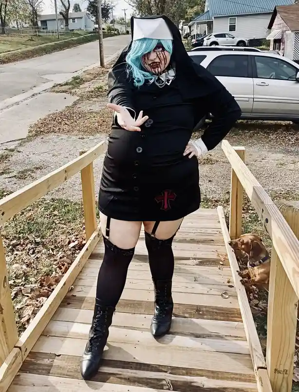 A person with blue hair and zombie makeup, dressed in a black nurse outfit with a red cross, stands on a wooden ramp. They…
