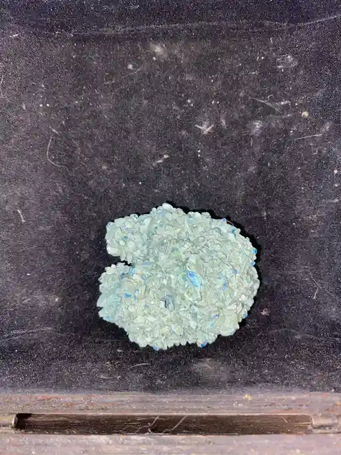 A close-up photo shows a small, irregularly shaped cluster of translucent, light blue-green crystals resting on a dark…
