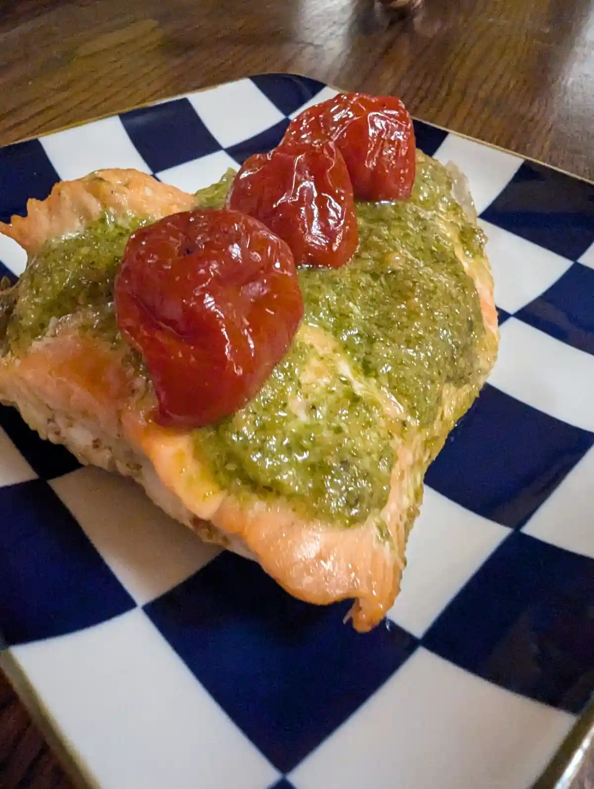 A close-up photo shows a cooked salmon fillet on a blue and white checkered plate, topped with green pesto sauce and three…