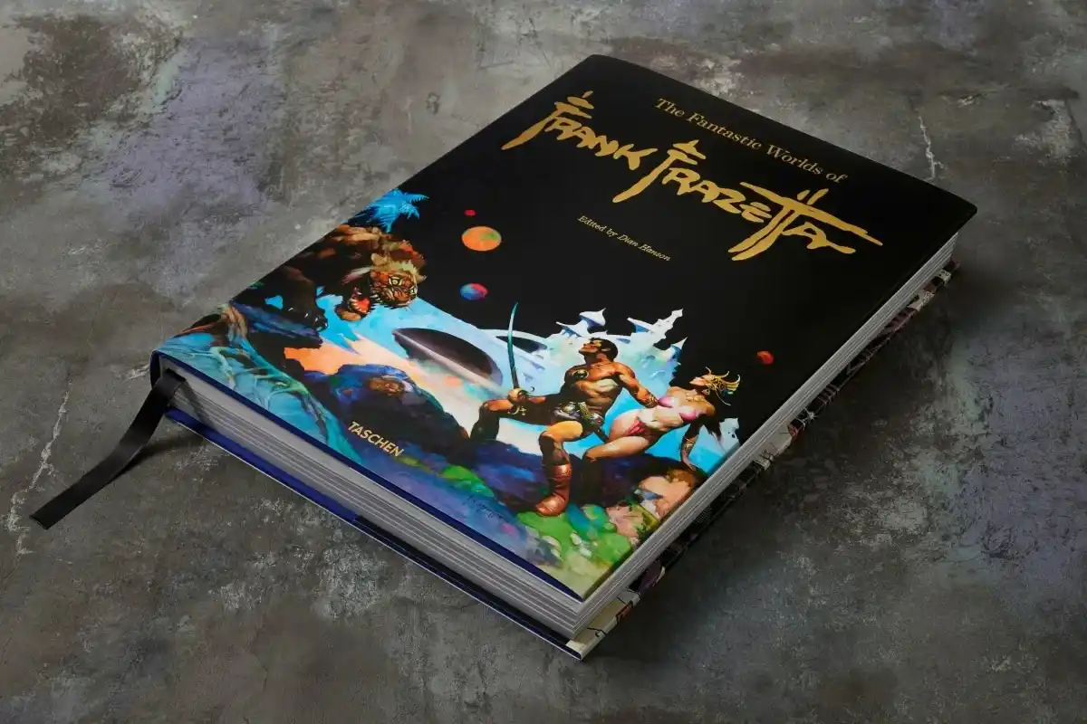 A large, hardcover art book titled 'The Fantastic Worlds of Frank Frazetta' lies on a textured gray surface. The cover…
