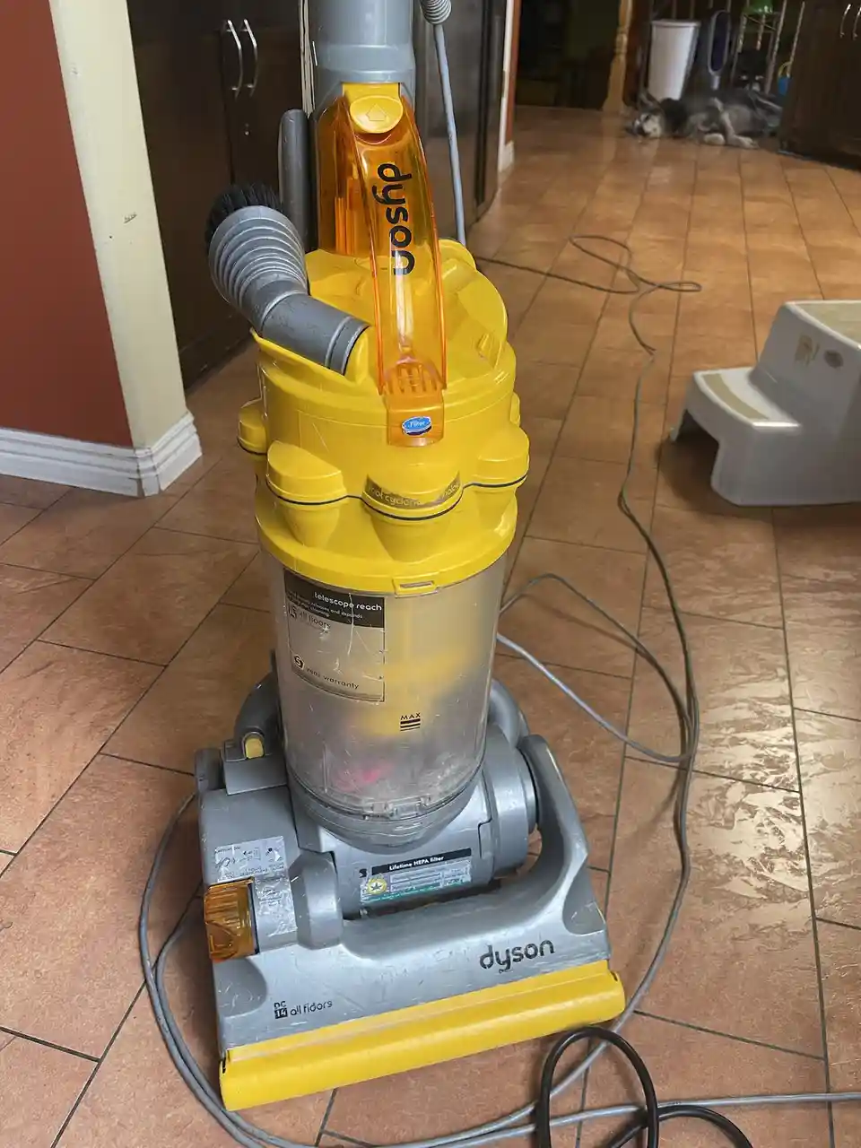 A yellow and gray Dyson vacuum cleaner sits on a tiled floor in a home, with a dog sleeping in the background. The vacuum…