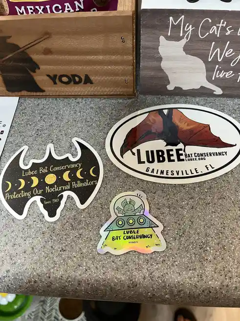 Three stickers from the Lubee Bat Conservancy are displayed on a speckled countertop, featuring a bat-shaped sticker with…