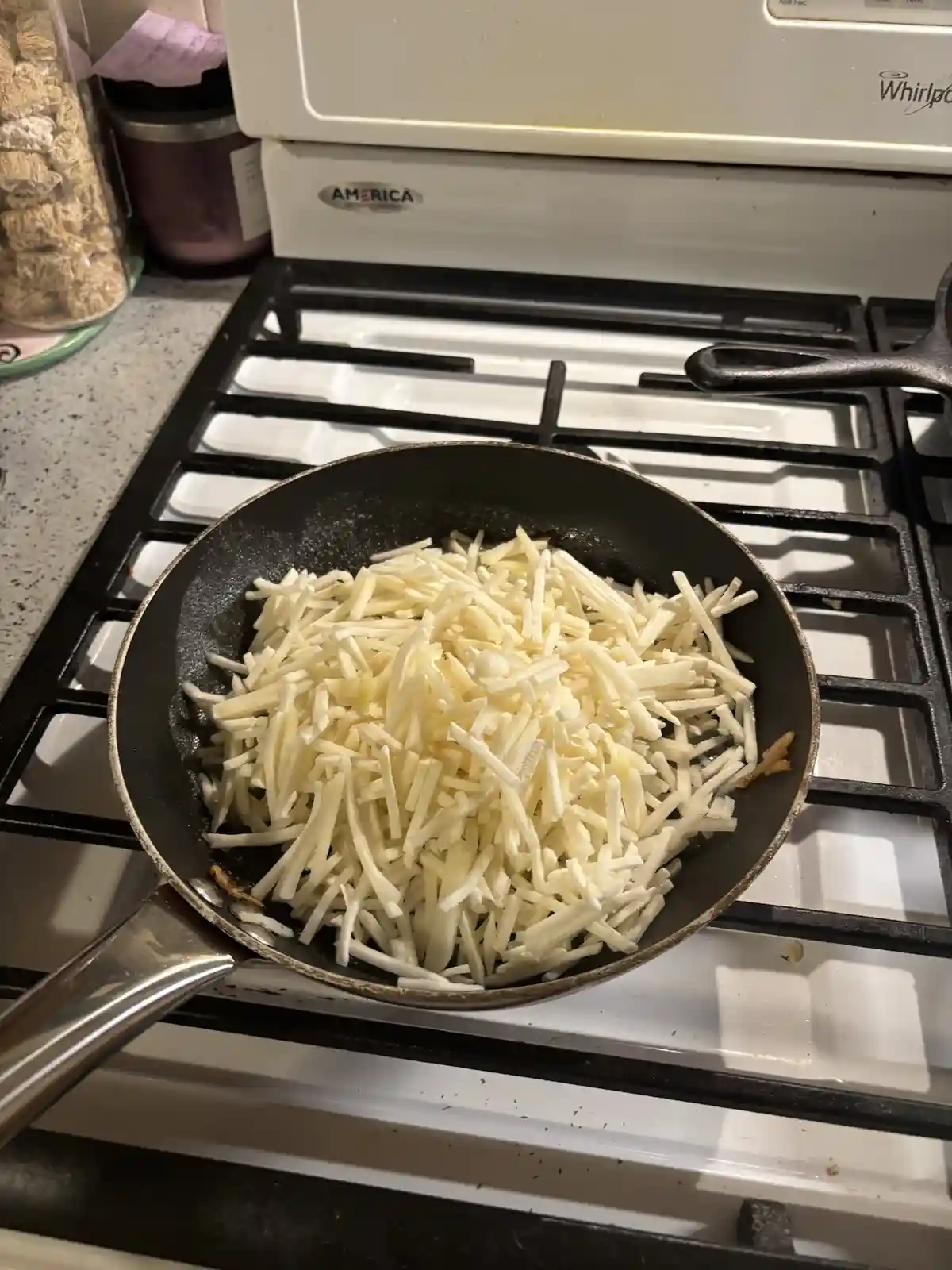 A black frying pan sits on a white gas stove burner, filled to the brim with a large mound of shredded white cheese. The pan…