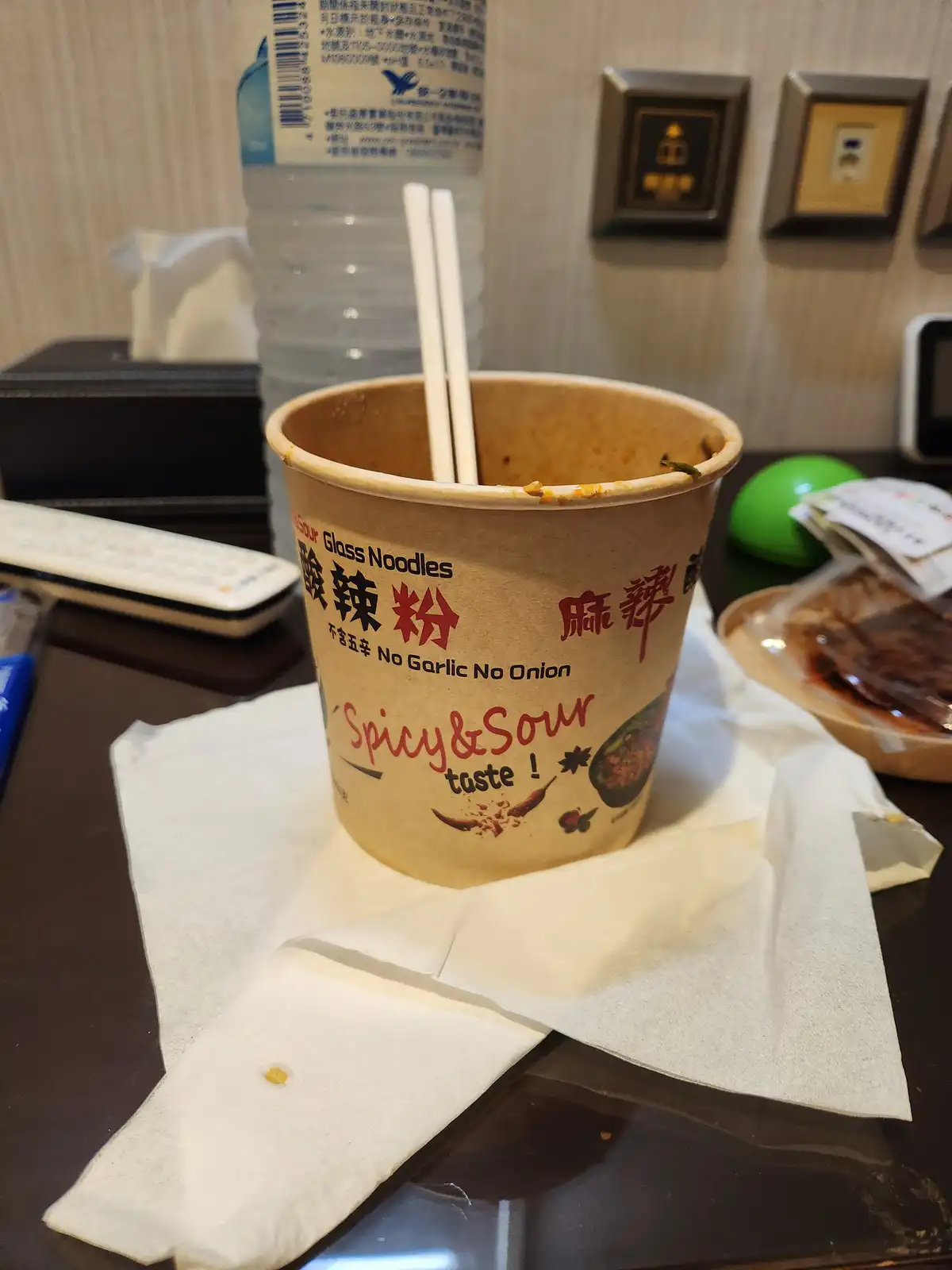 An empty, stained cup of spicy and sour glass noodles sits on a napkin on a hotel room desk, with chopsticks inside and a…