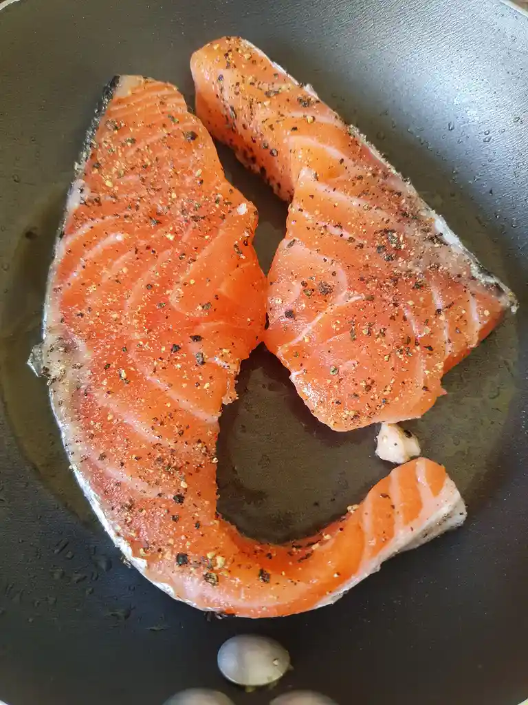 Two raw salmon fillets seasoned with black pepper and other spices are sizzling in a dark non-stick frying pan, with oil…