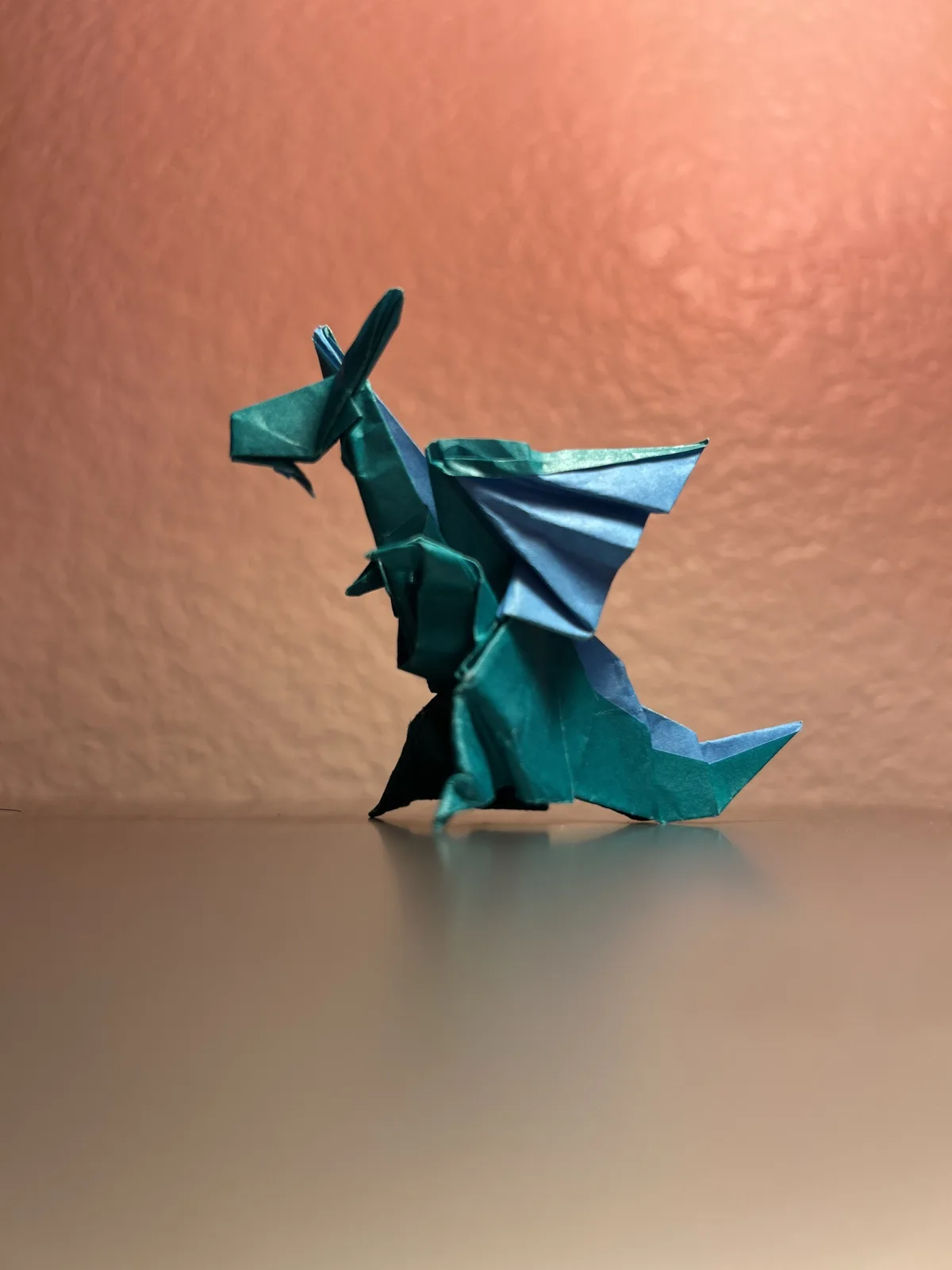 A teal and blue origami dragon stands on a reflective surface against a textured peach-colored wall. The dragon is made from…