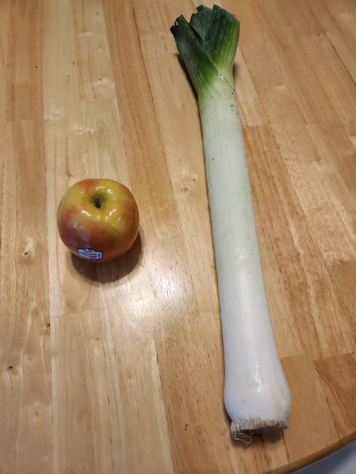 A red and yellow apple with a sticker sits next to a long green and white leek on a light wooden surface. The overlaid text…