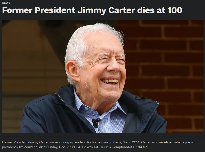 A smiling, elderly Jimmy Carter with white hair, wearing a dark jacket over a checkered shirt, is pictured against a brick…