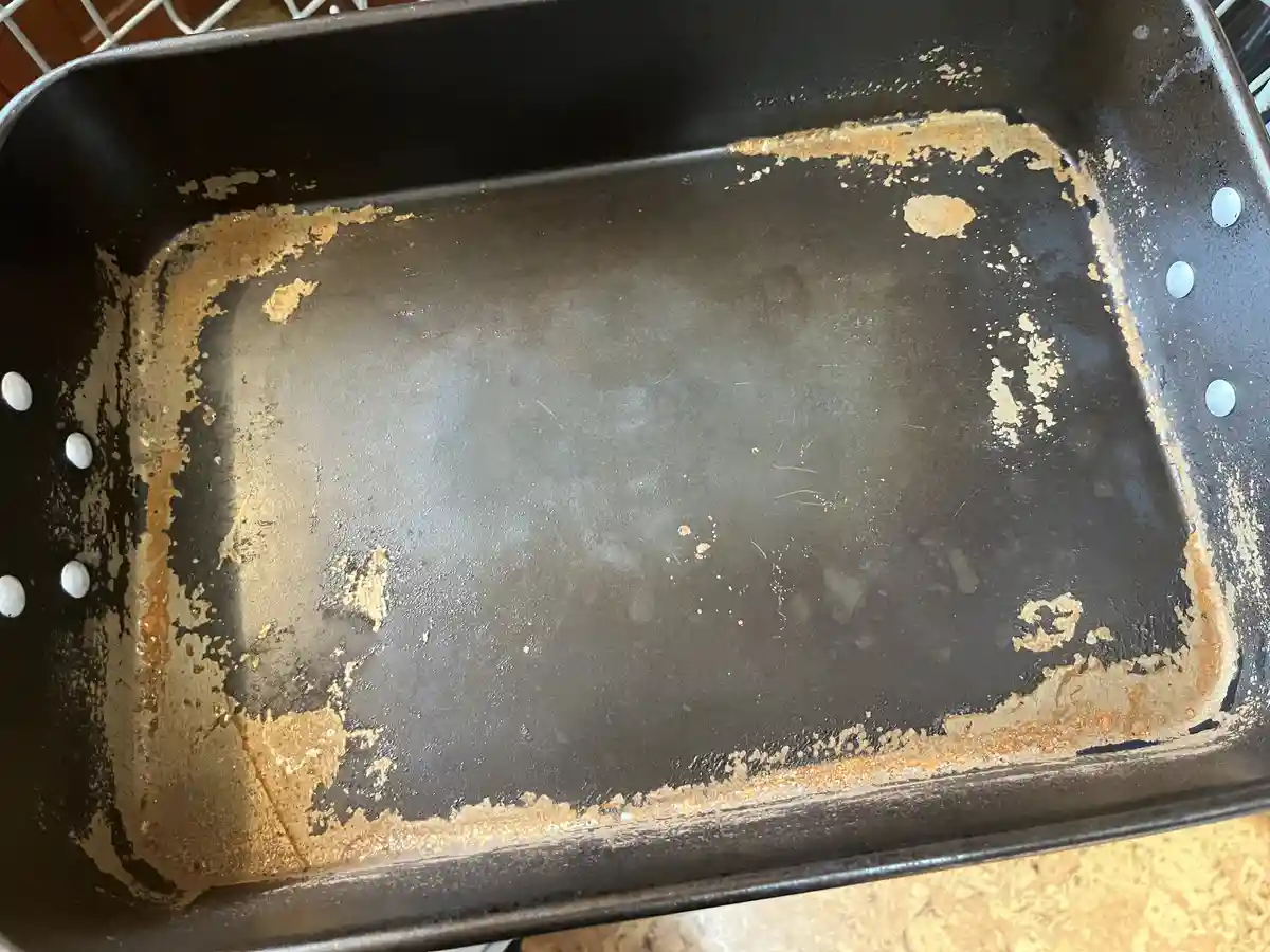 a close-up photo of a heavily stained, rusted, and worn black metal baking pan with orange-brown residue along its edges and…