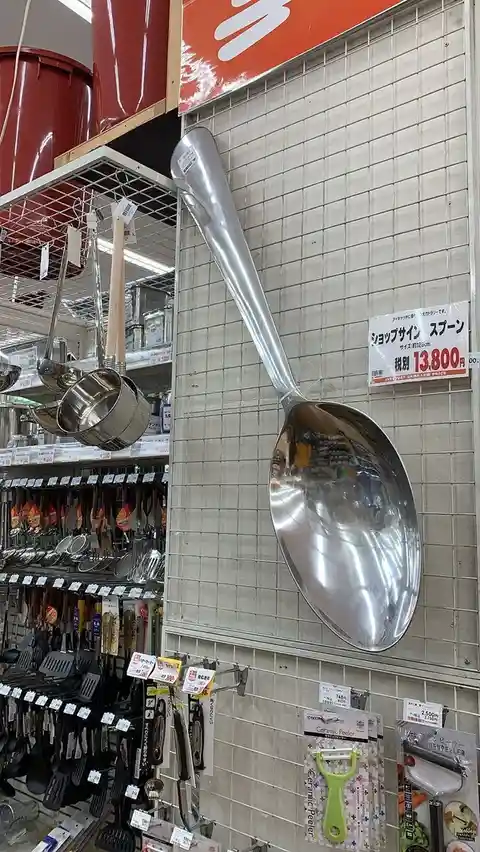 A giant, shiny, metallic serving spoon, approximately 125cm long, is mounted on a tiled wall in a Japanese kitchenware…