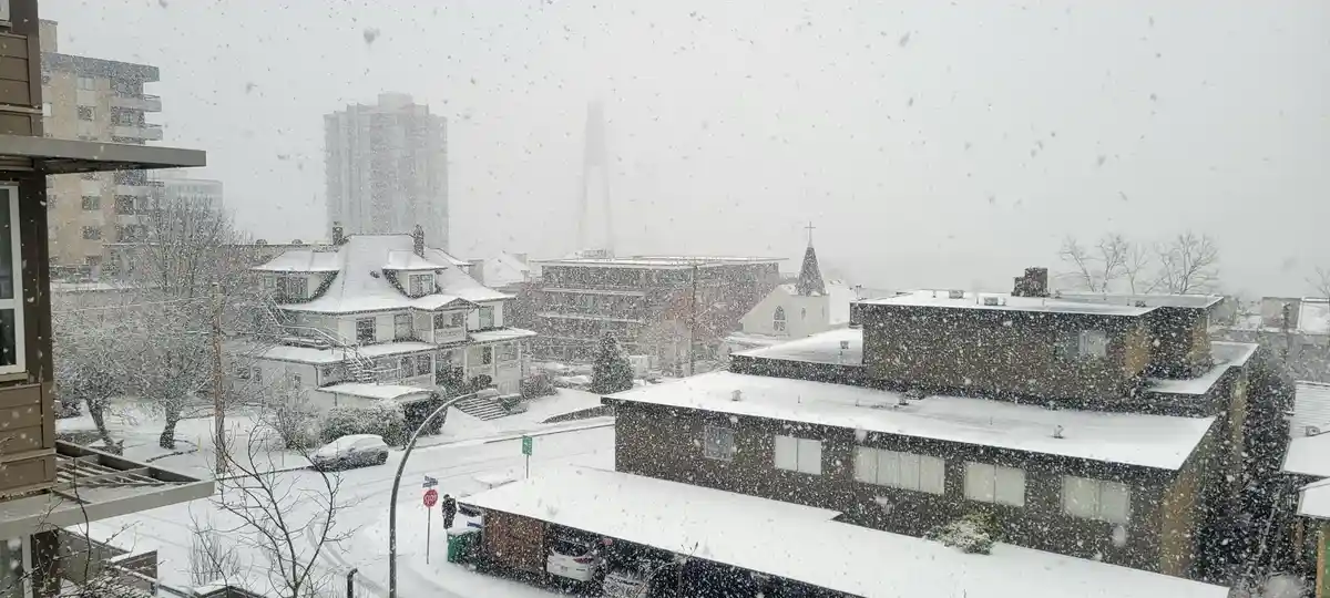 A heavy snowfall blankets a cityscape with rooftops, trees, and streets covered in white. Buildings of various styles are…