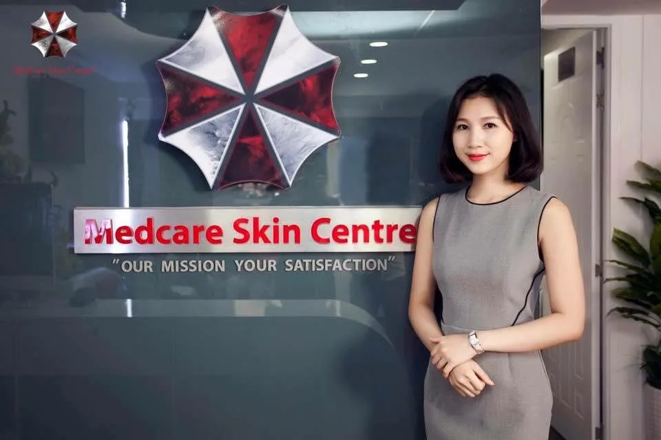 A woman in a grey dress stands smiling in front of a glass wall displaying a sign for 'Medcare Skin Centre' with the slogan…
