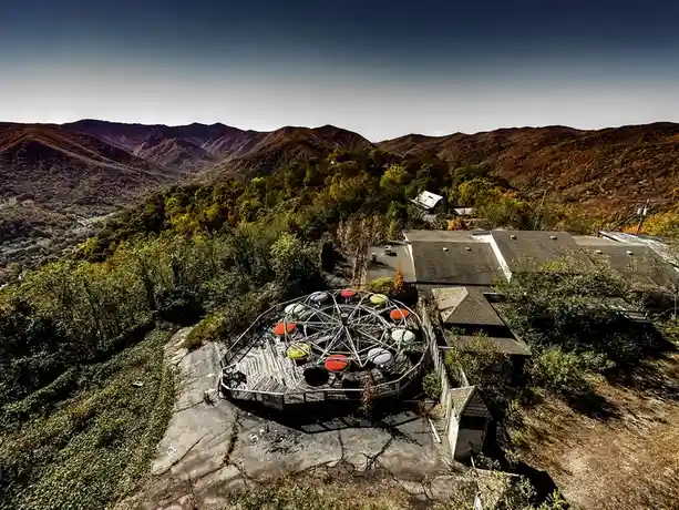 An aerial view shows a dilapidated, circular carnival ride with colorful seats, labeled 'CATCHER', sitting on cracked…