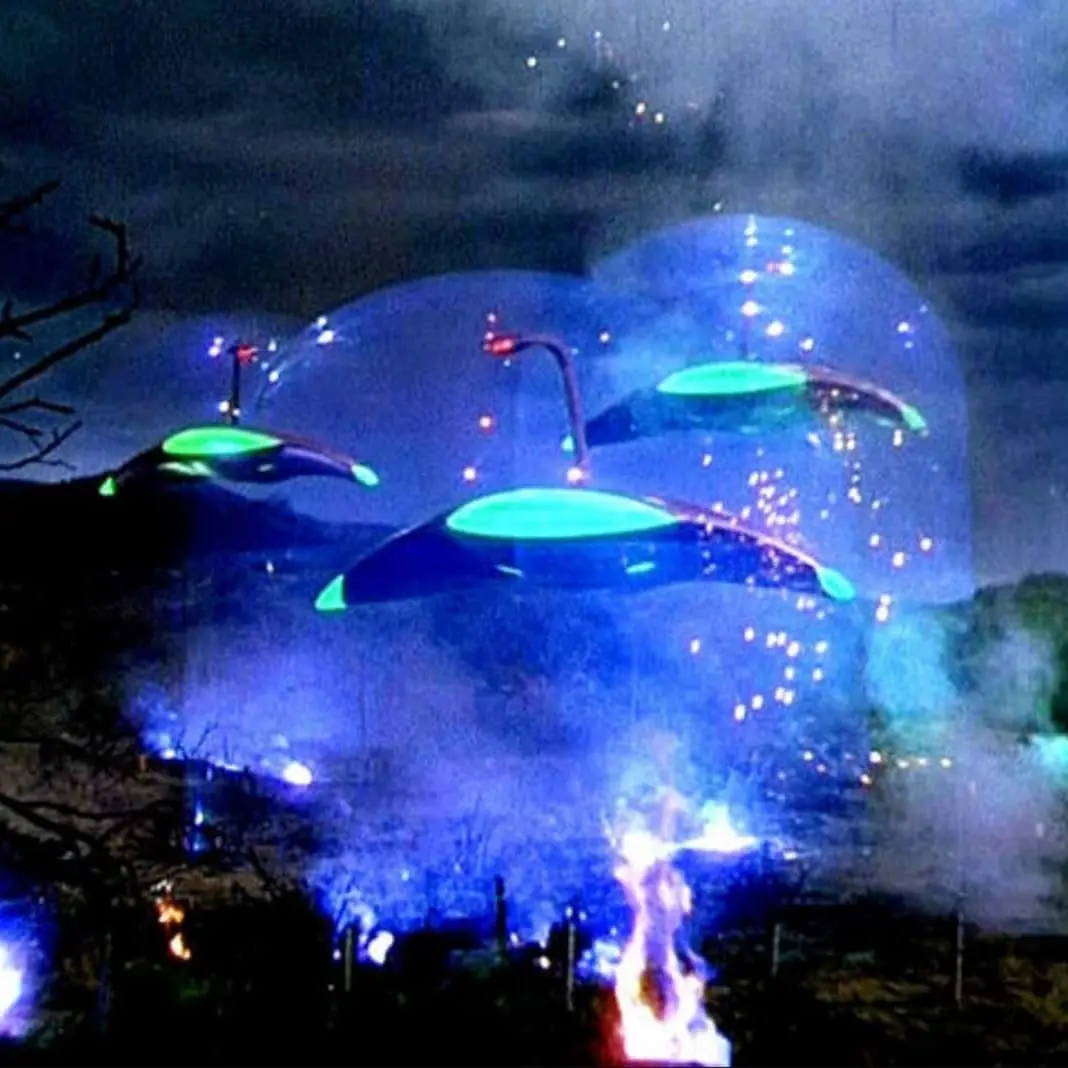 A nighttime scene shows several glowing, saucer-shaped UFOs hovering over a landscape with trees and a visible fire burning…